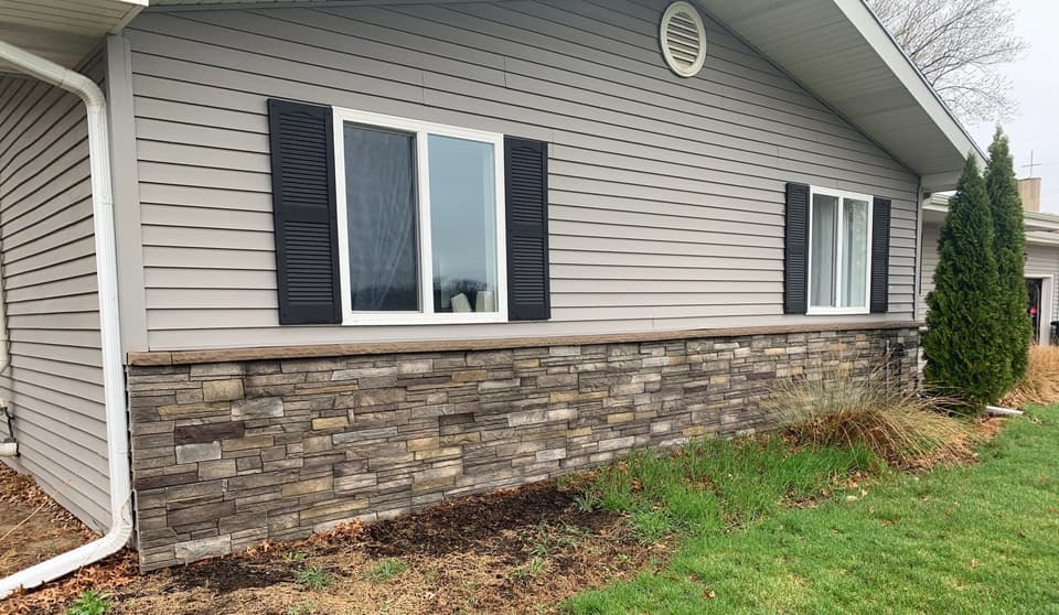 A mobile home foundation has been retrofitted with a stone veneer skirt that covers the lower exterior, with new windows and black shutters visible above, showing completed foundation restoration work.
