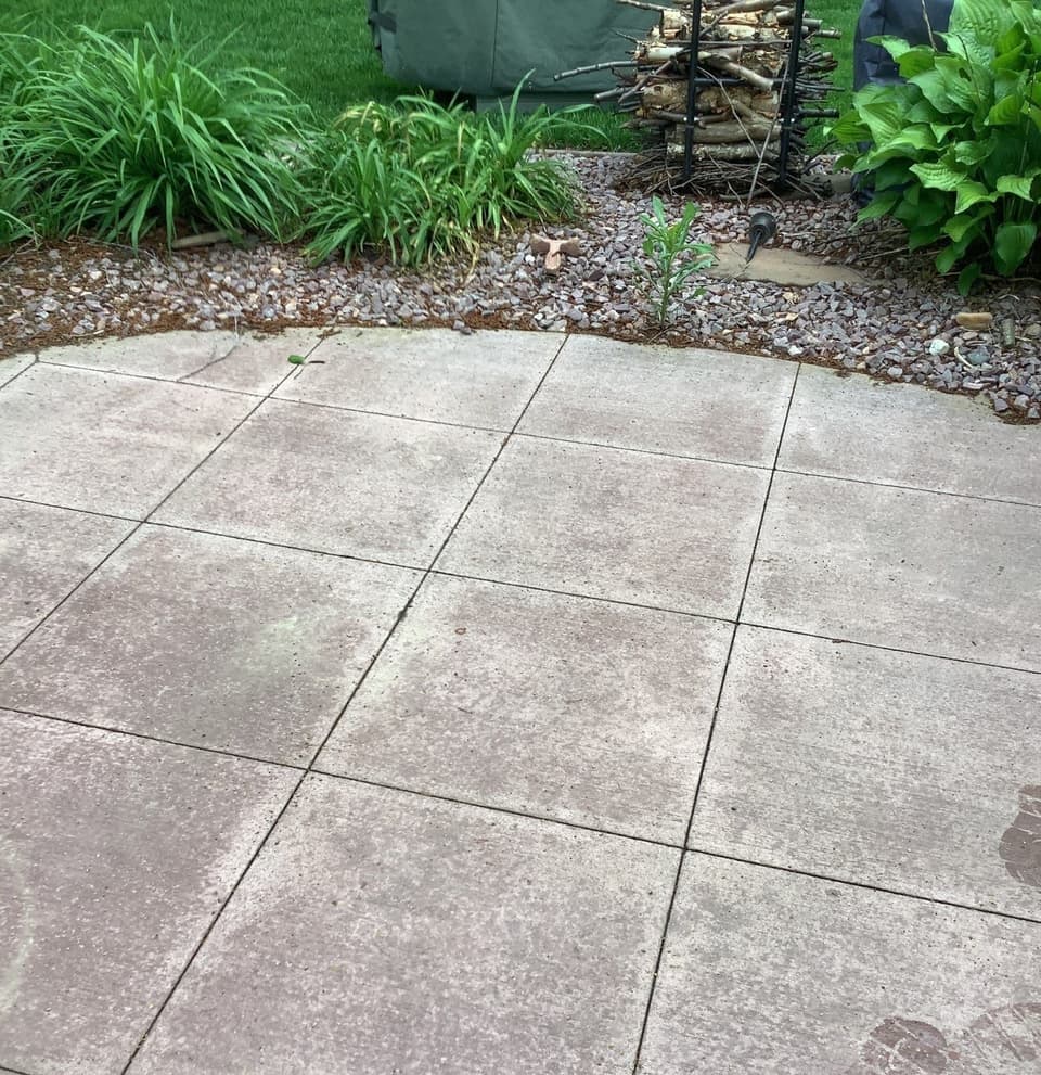 Completed concrete patio or foundation flatwork with uniform tile pavers laid in a grid pattern, bordered by landscaping and gravel.