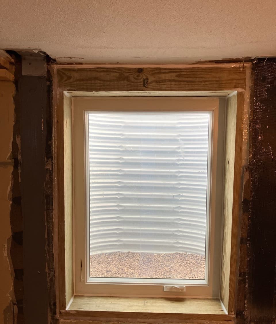 A newly installed egress window with light wood frame and sill looking out to a beach with water and sand visible through the glass.