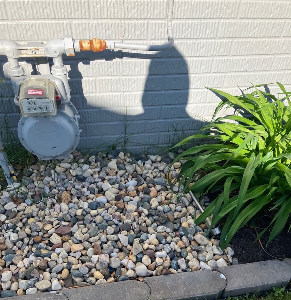 A completed sump pump installation with drainage system visible against a brick foundation, surrounded by decorative river rocks and landscaping edging.