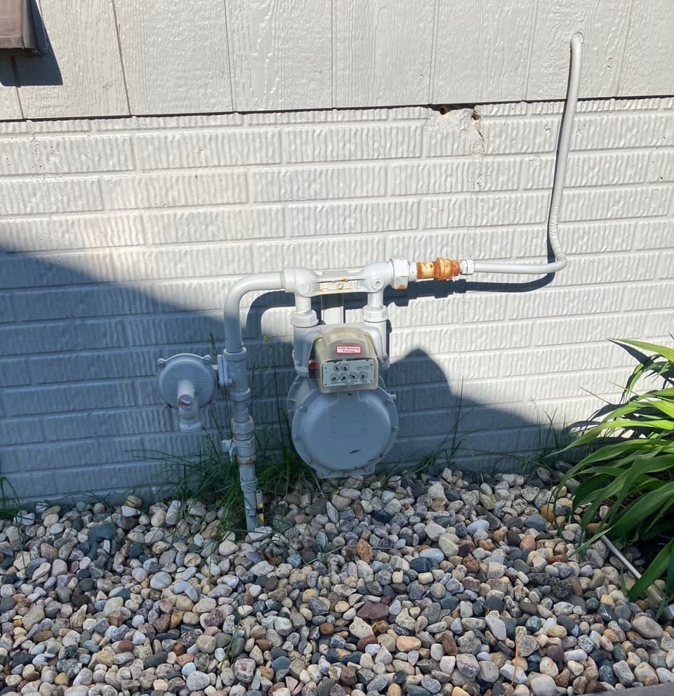 A completed sump pump installation system is visible against a brick foundation with decorative rock bed and landscaping, showing finished installation with discharge piping.