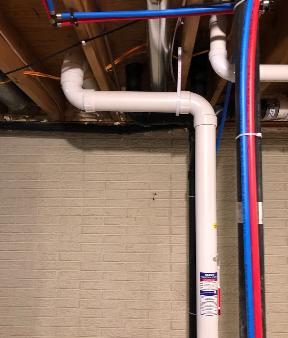 Installed sump pump system with white PVC discharge pipe and integrated interior drainage system alongside blue and red PEX tubing in a basement setting.