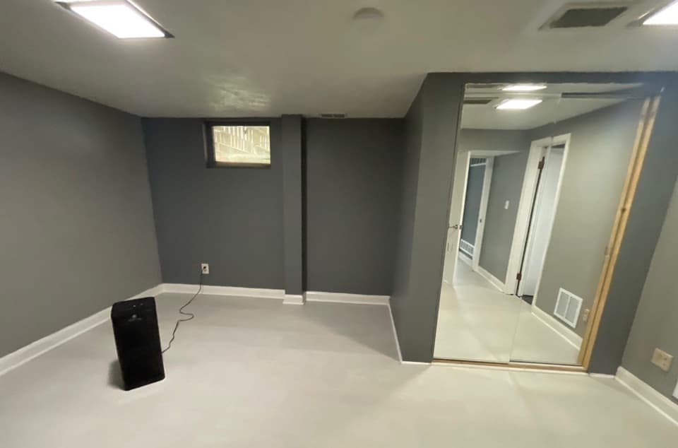 A finished basement or crawlspace renovation showing new white epoxy-coated flooring, gray painted walls, recessed lighting, and what appears to be a dehumidifier unit in place.