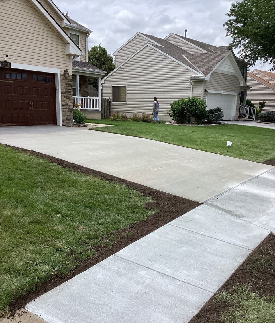 Newly poured concrete walkway and driveway with fresh concrete flatwork visible alongside freshly prepared landscaping beds with dark mulch and seeded lawn areas.