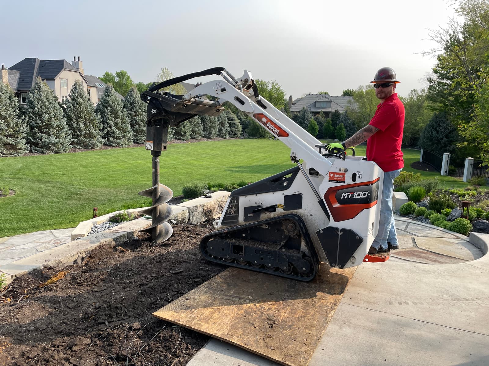 Worker operating a Bobcat mini skid steer with an auger attachment, drilling into the ground for helical piers.