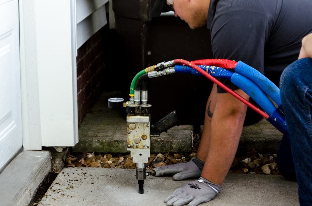 Worker injecting polyurethane foam to lift a concrete slab next to a building foundation.