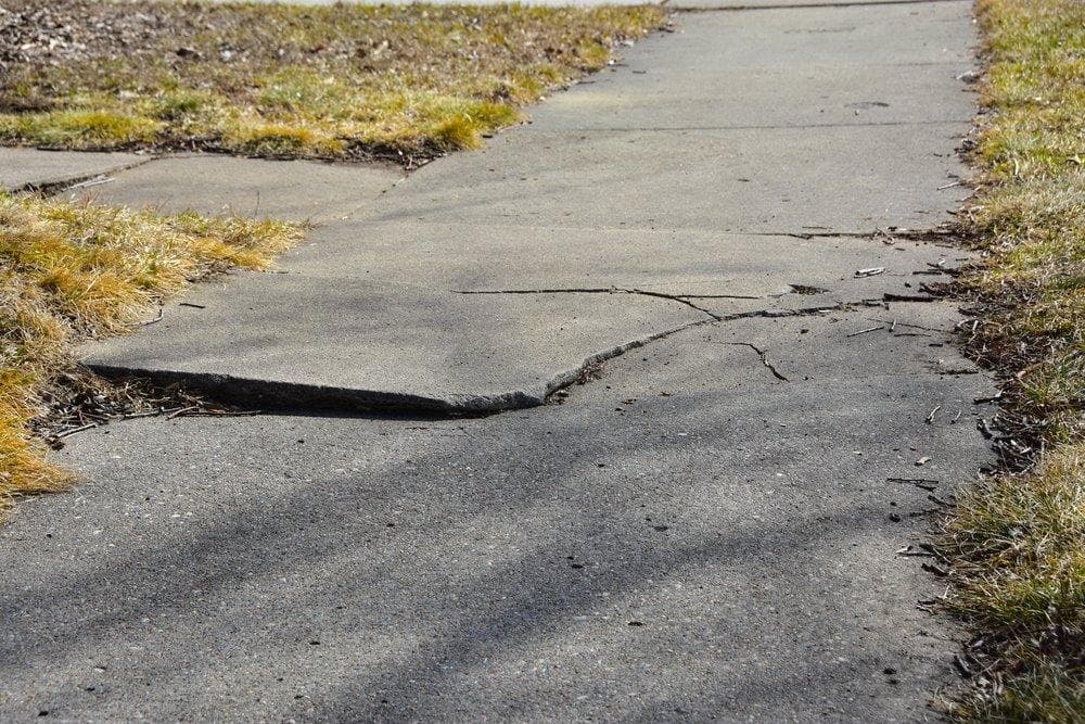 Cracked and uneven concrete sidewalk with signs of soil settlement.