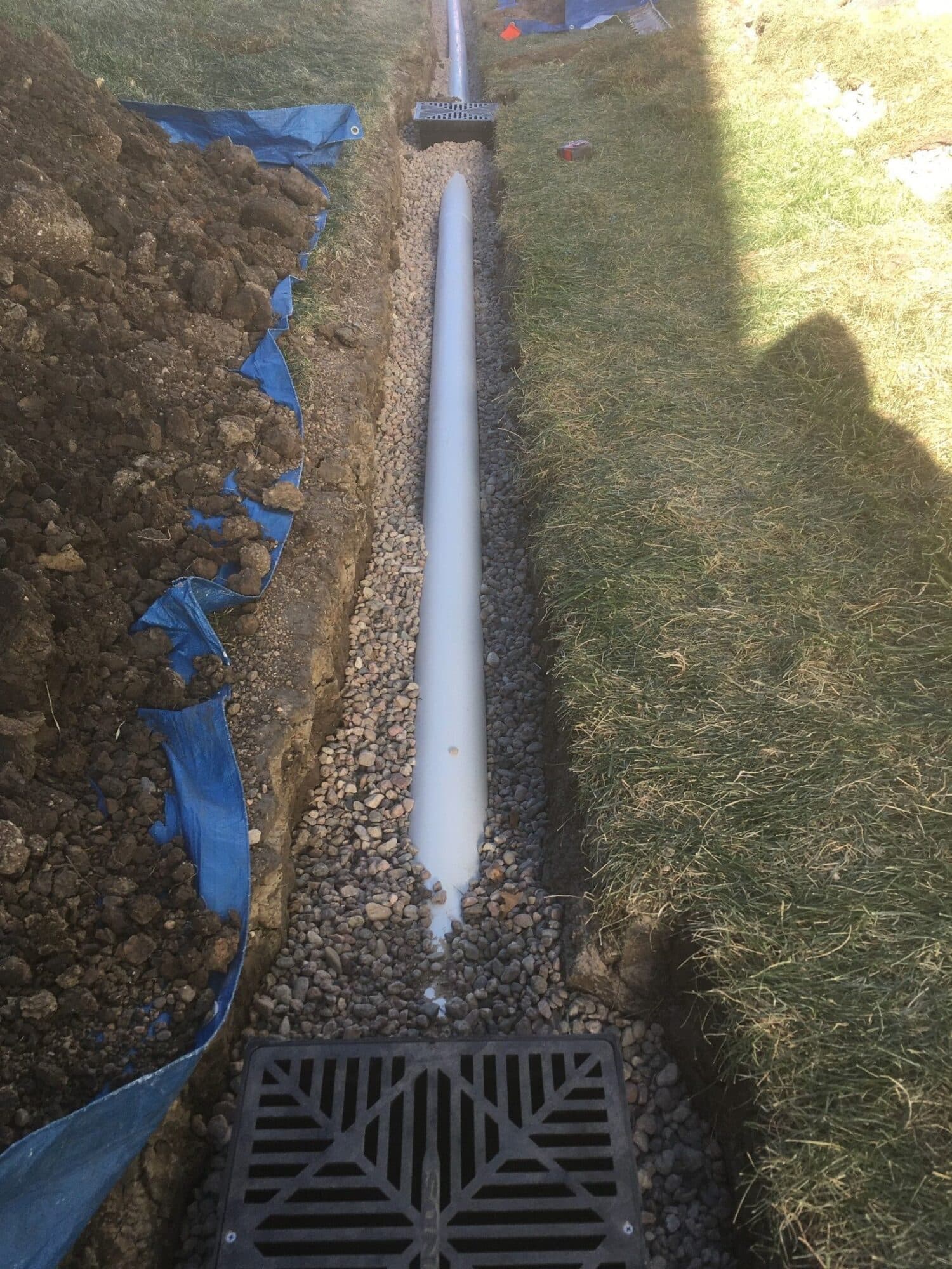 Overhead photo of an open yard drainage trench with white pipe gravel backfill and a catch basin grate
