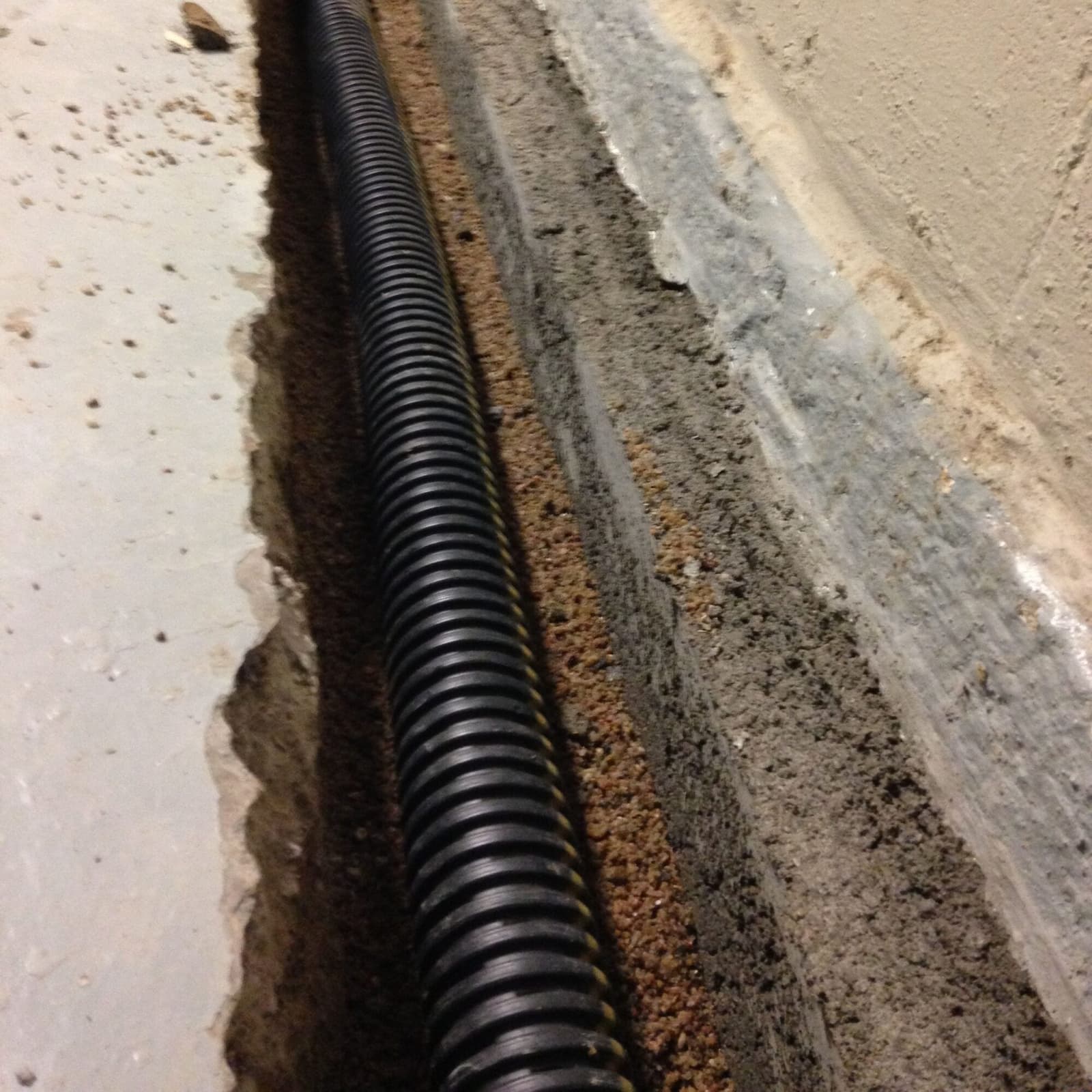 Close up real photo of a black corrugated drain pipe laid in a gravel trench along the base of a basement wall