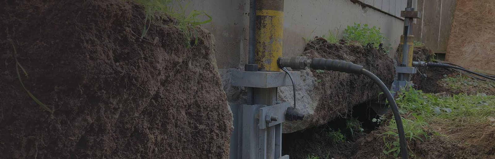 Wide hero background photo of hydraulic pier equipment and a drainage hose at an excavated foundation wall