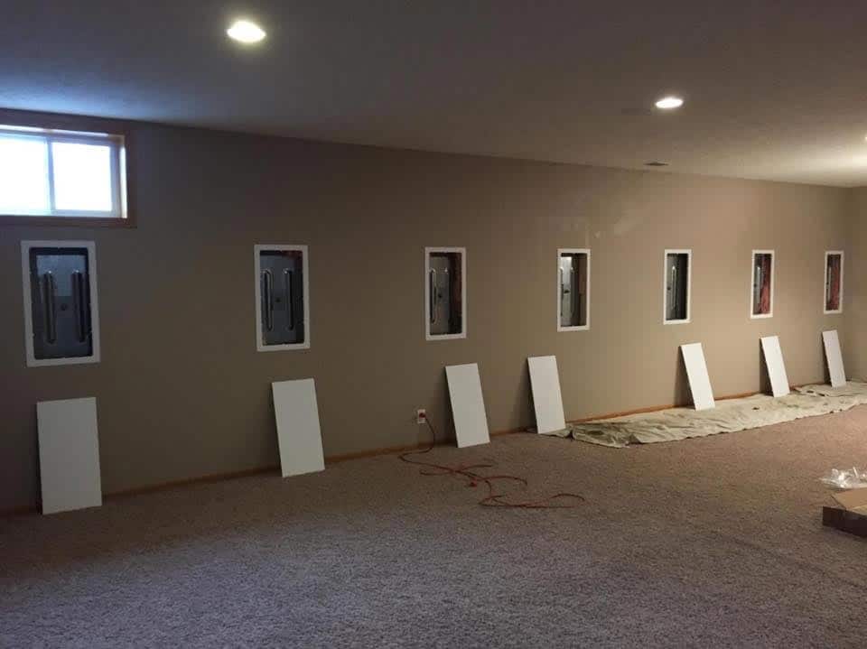 Real photo of a finished basement with several rectangular steel brace access panels installed along a painted wall and additional white panels leaning against the wall ready to install